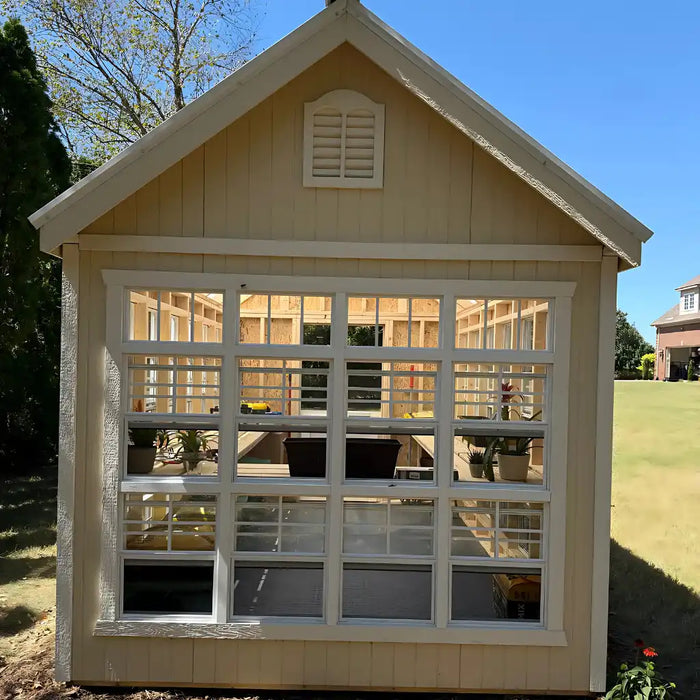 Small beige colonial gable shed greenhouse with white window frame in a garden setting