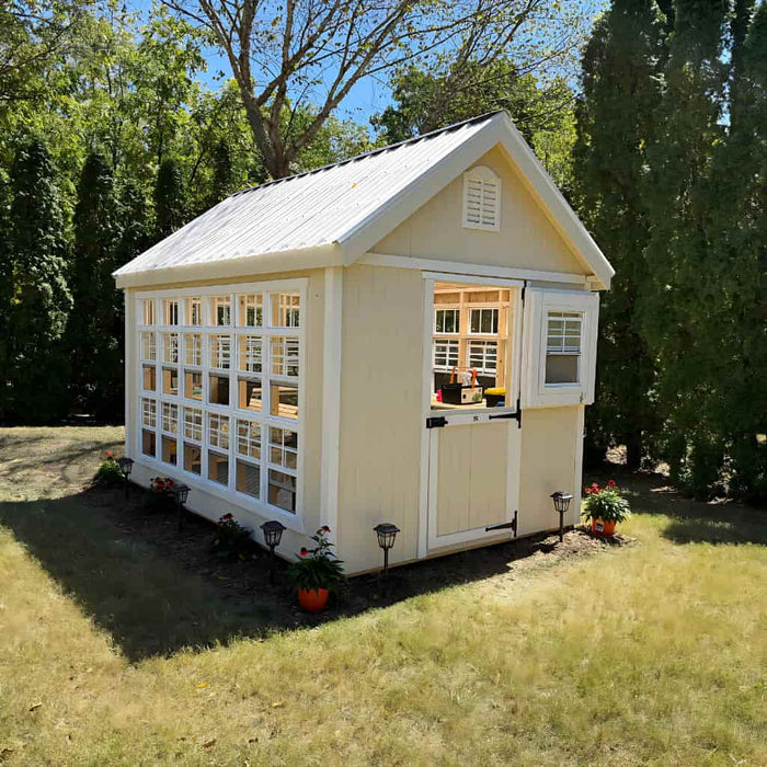 Small beige colonial gable greenhouse with white trim in a garden setting