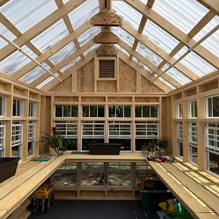 Colonial gable greenhouse interior with transparent roof and walls, featuring workbenches and tools.