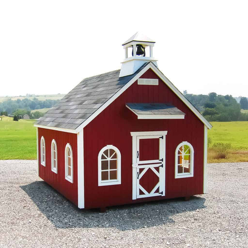 Little Cottage Co Stratford Schoolhouse playhouse kit with white trim on a gravel surface with a grassy field and trees in the background.