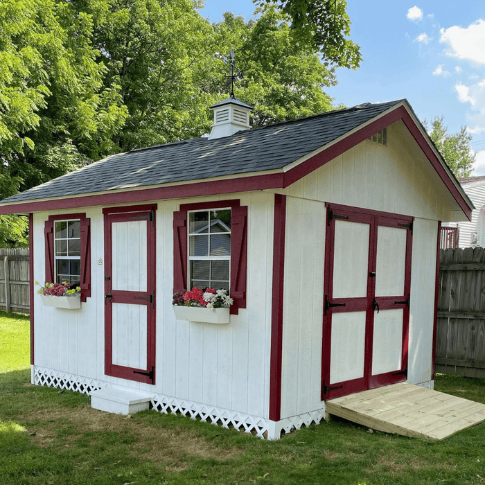 White and red trim garden shed with double doors and side windows, set against a lush backyard setting.